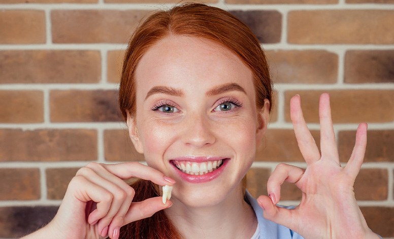 Woman with red hair holding extracted tooth and making “ok” sign with other hand.