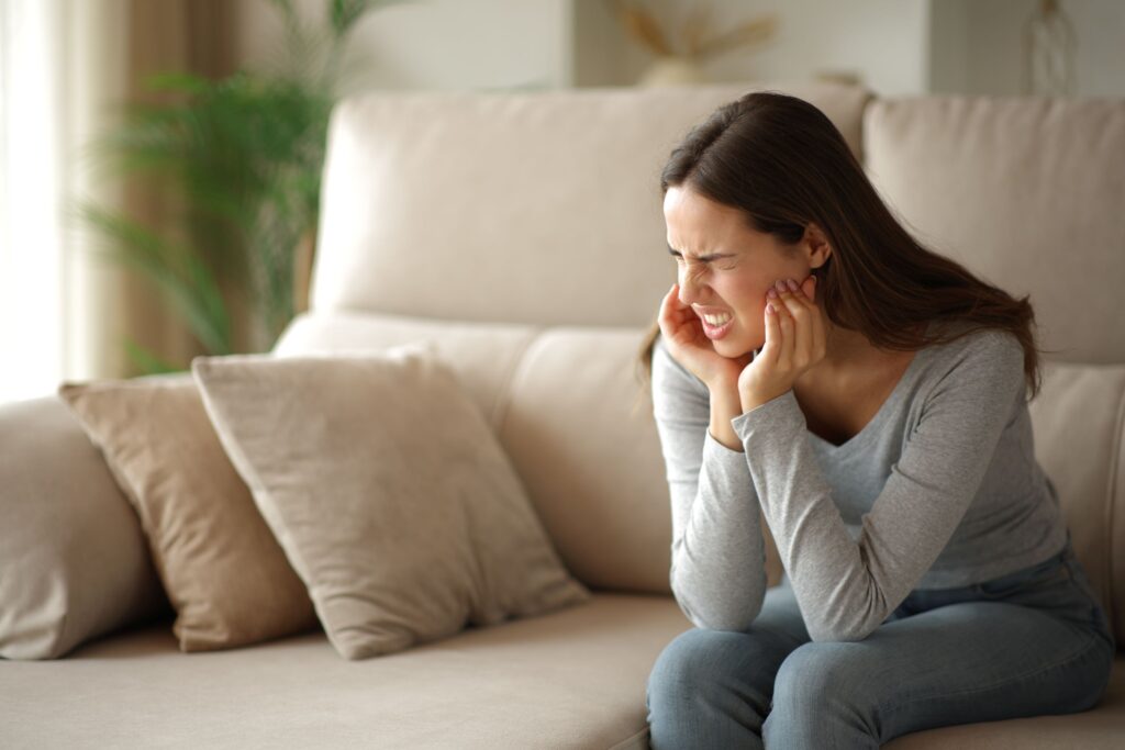 Woman on beige couch squinting and touching jaw in pain
